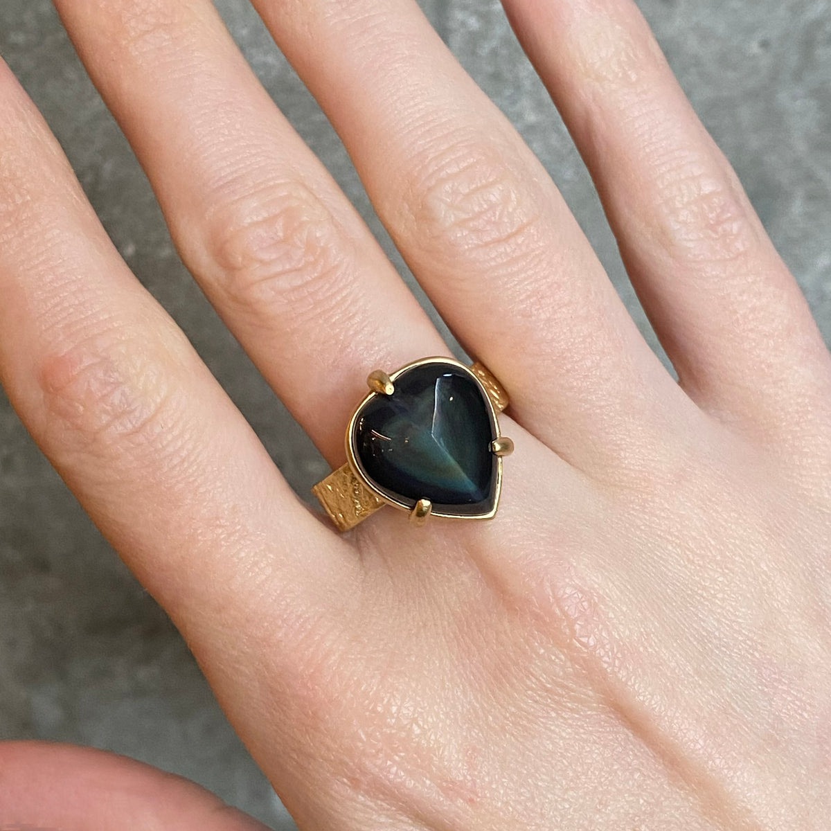 A gold ring with a heart-shaped dark stone featuring a rainbow cats-eye play-of-colour, worn on a person's hand. The band is wide textured gold.