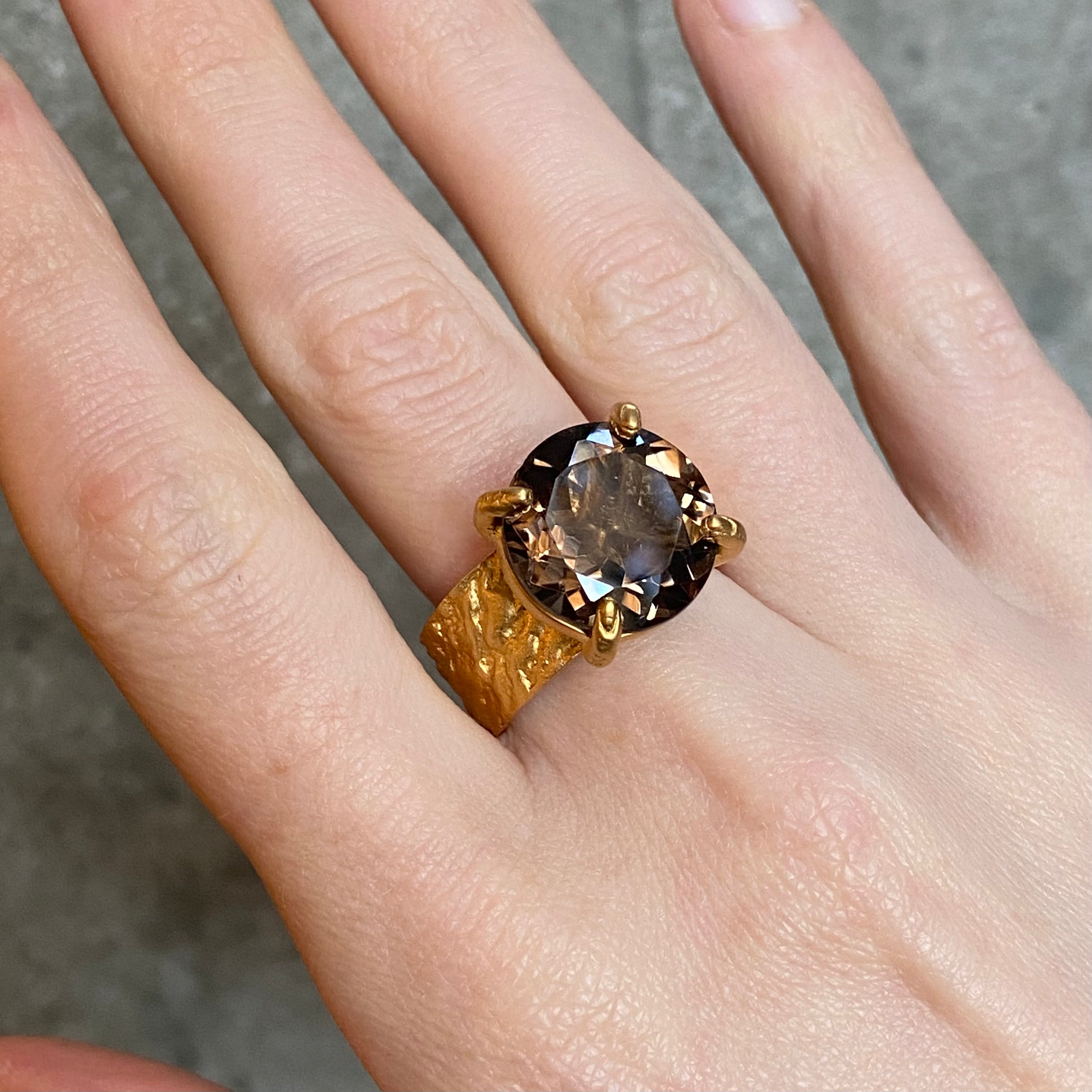 A gold cocktail ring with a large round brown gemstone on a wide textured band, set with four claws, worn on a person's finger against a neutral grey concrete background.