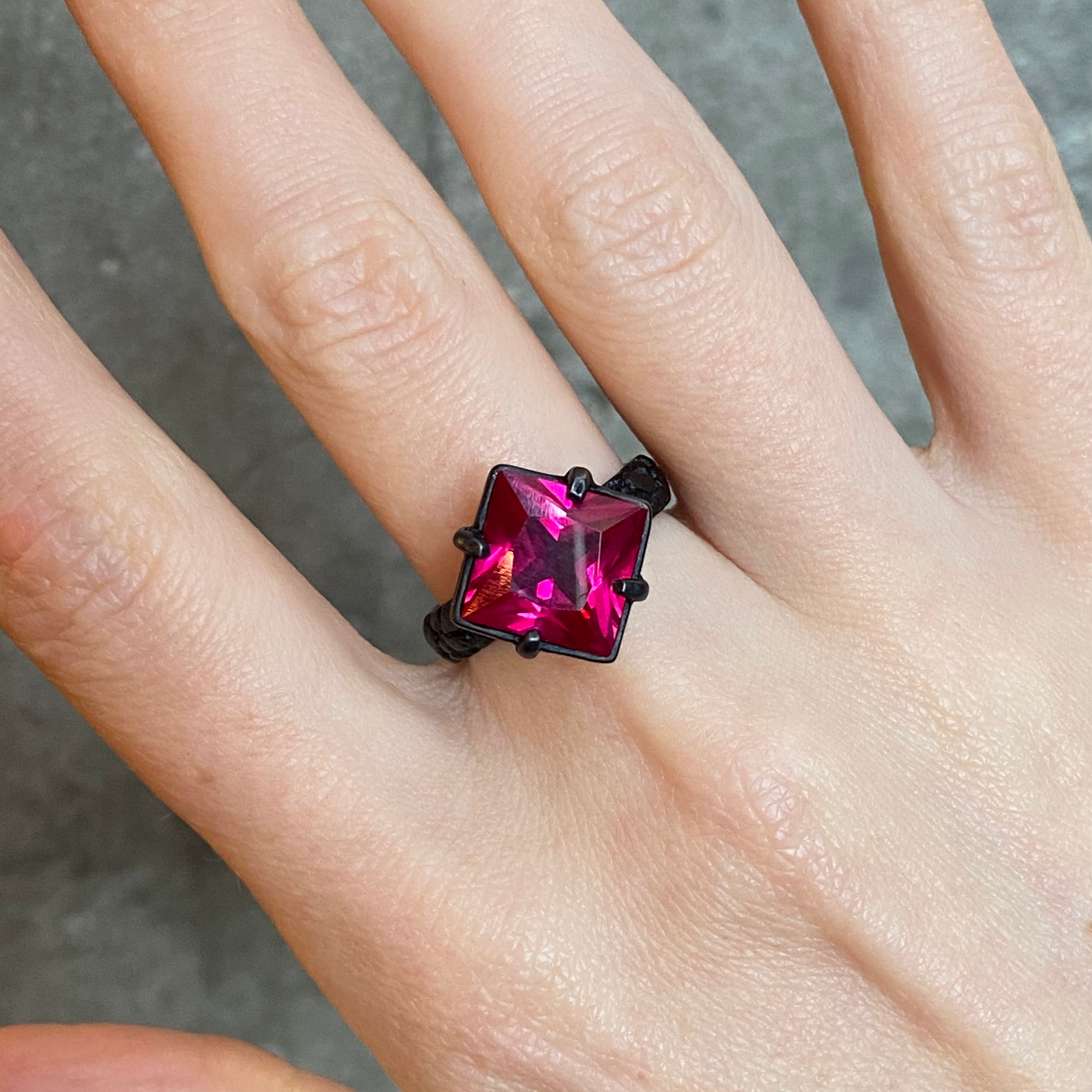 A close-up of a hand wearing a black ring with a large, square-cut, vivid magenta-pink gemstone set in four claws.