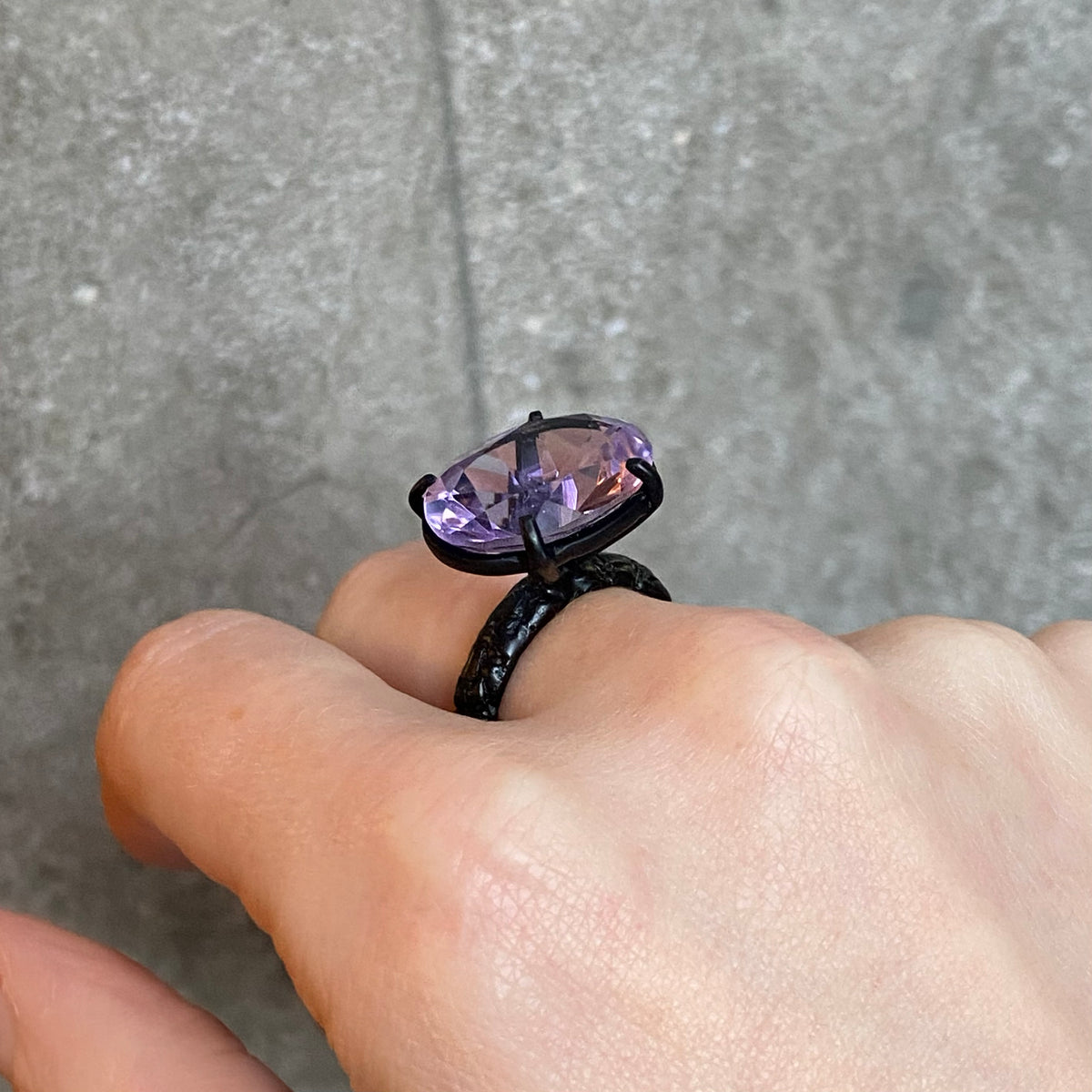 A close-up of a hand wearing a large black cocktail ring with a large faceted purple gemstone held by four black claws, set against a grey textured background.
