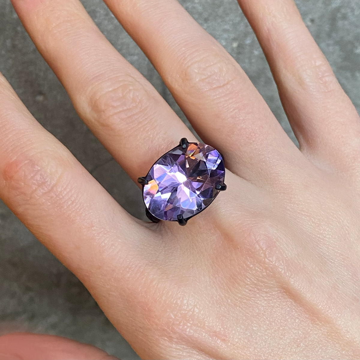 A close-up of a hand wearing a ring with a large faceted oval purple gemstone in a black metal four claw setting, against a grey concrete background.
