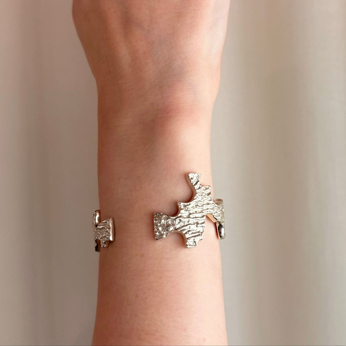 A close-up of a person's wrist wearing an organic textured silver cuff bracelet with irregular cut-out edges, set against a soft beige curtain background.