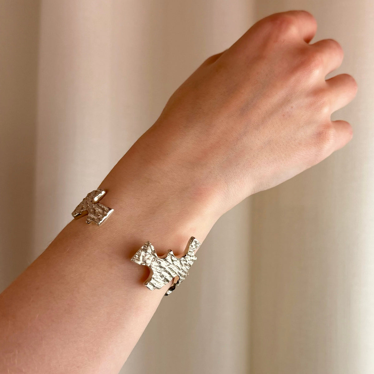 A close-up of a person's wrist wearing an organic textured silver cuff bracelet with irregular cut-out edges, set against a soft beige curtain background.