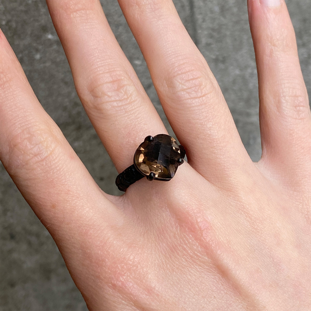 A close up of a hand wearing a ring with a large faceted smoky brown gemstone in a heart shape on a black band, shown against a grey concrete background.