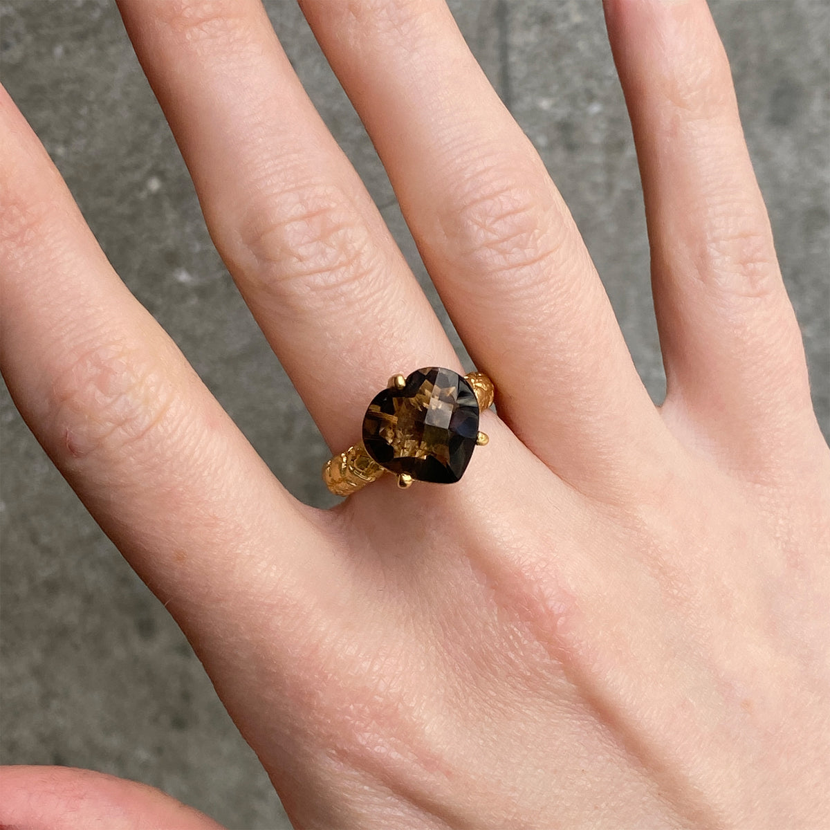 A gold ring with a faceted dark brown smoky gemstone worn on a person's finger, shown against a neutral gray concrete background.