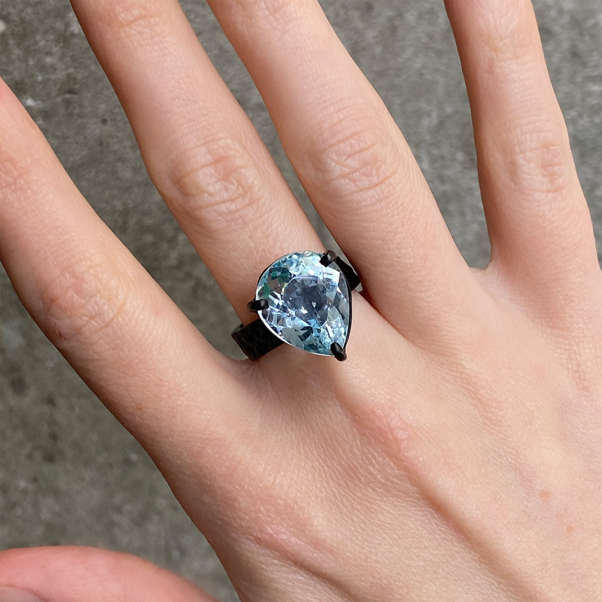 A close up of a hand wearing a black ring with a large pear cut light blue gemstone.