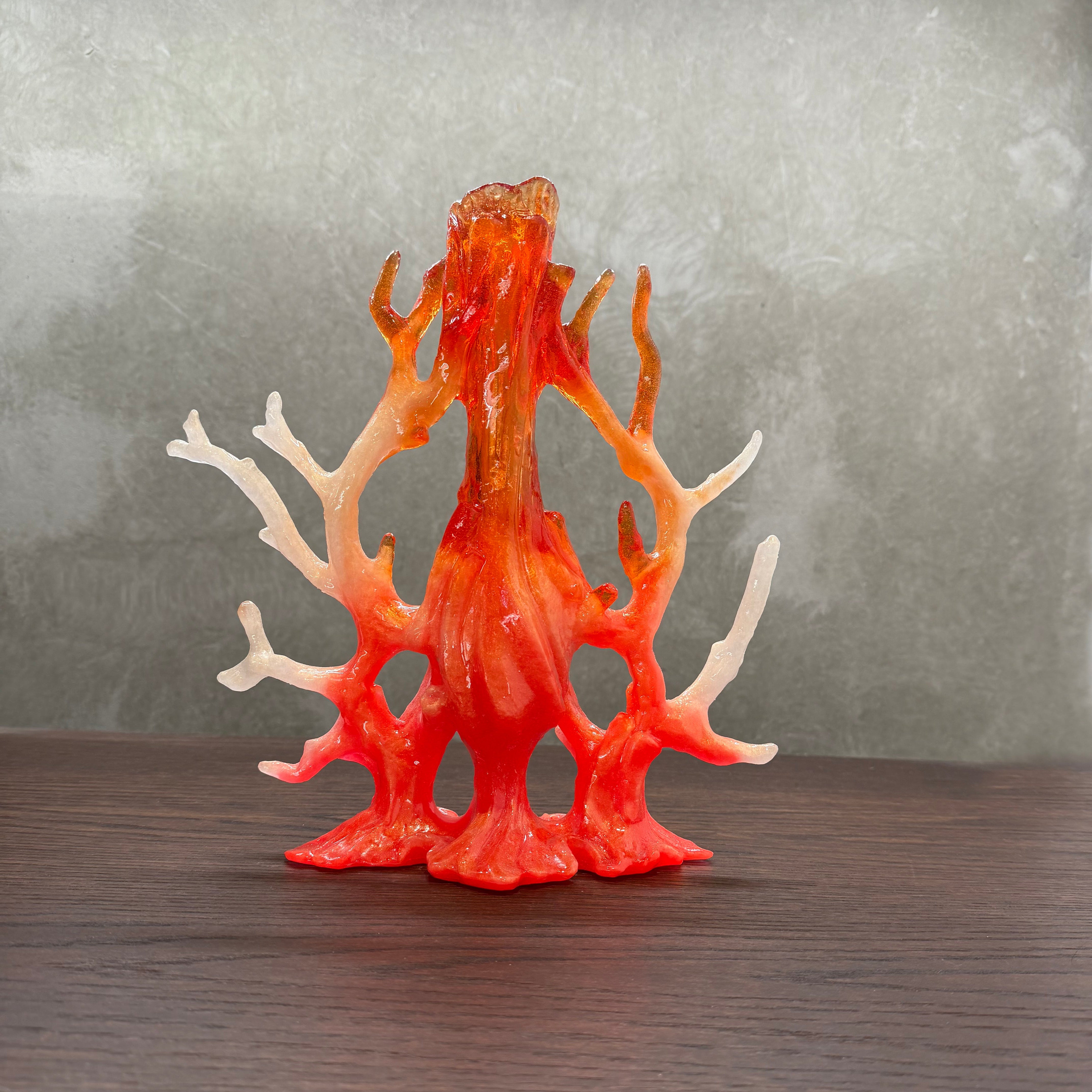 A coral shaped decorative sculptural vessel n a translucent gradient of orange, red, and cream, displayed on a dark wooden surface against a textured gray background.