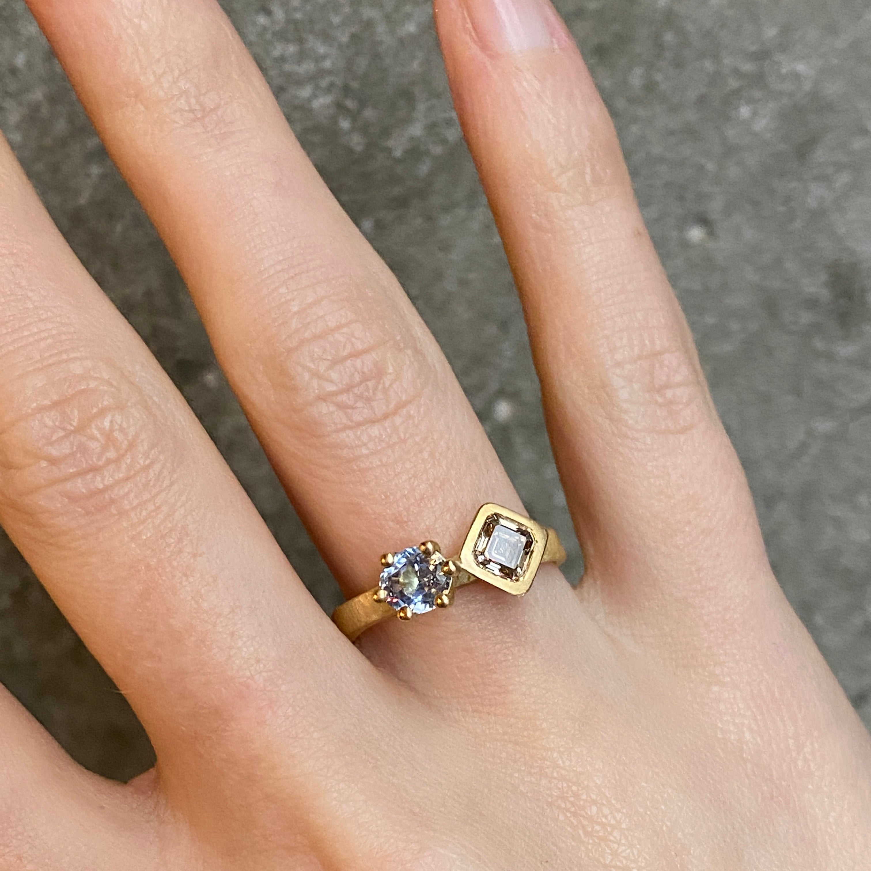 A gold ring with two gemstones of differnt shapes worn on a person's finger, photographed against a textured gray concrete background.