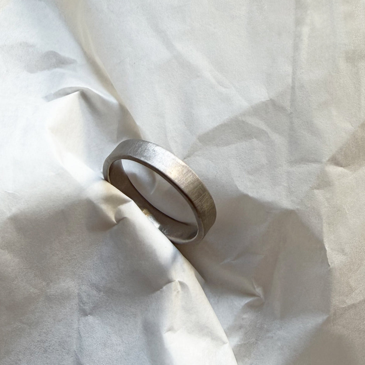A minimalist brushed white gold band ring resting on crumpled white paper.
