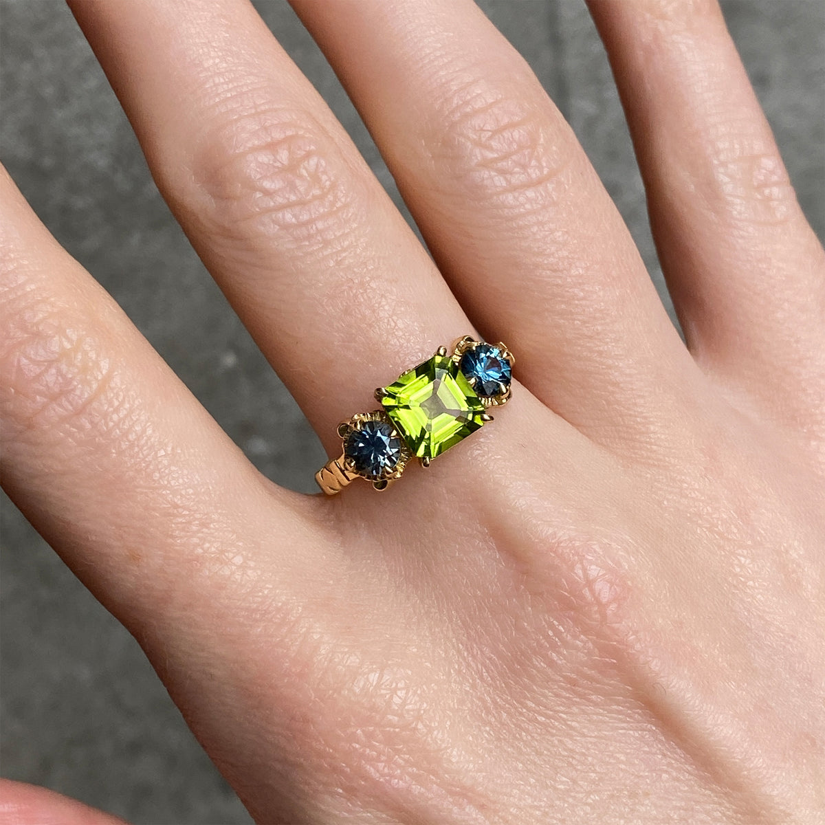 A gold ring with a central peridot and two round blue sapphires on the each side worn on a person's finger.
