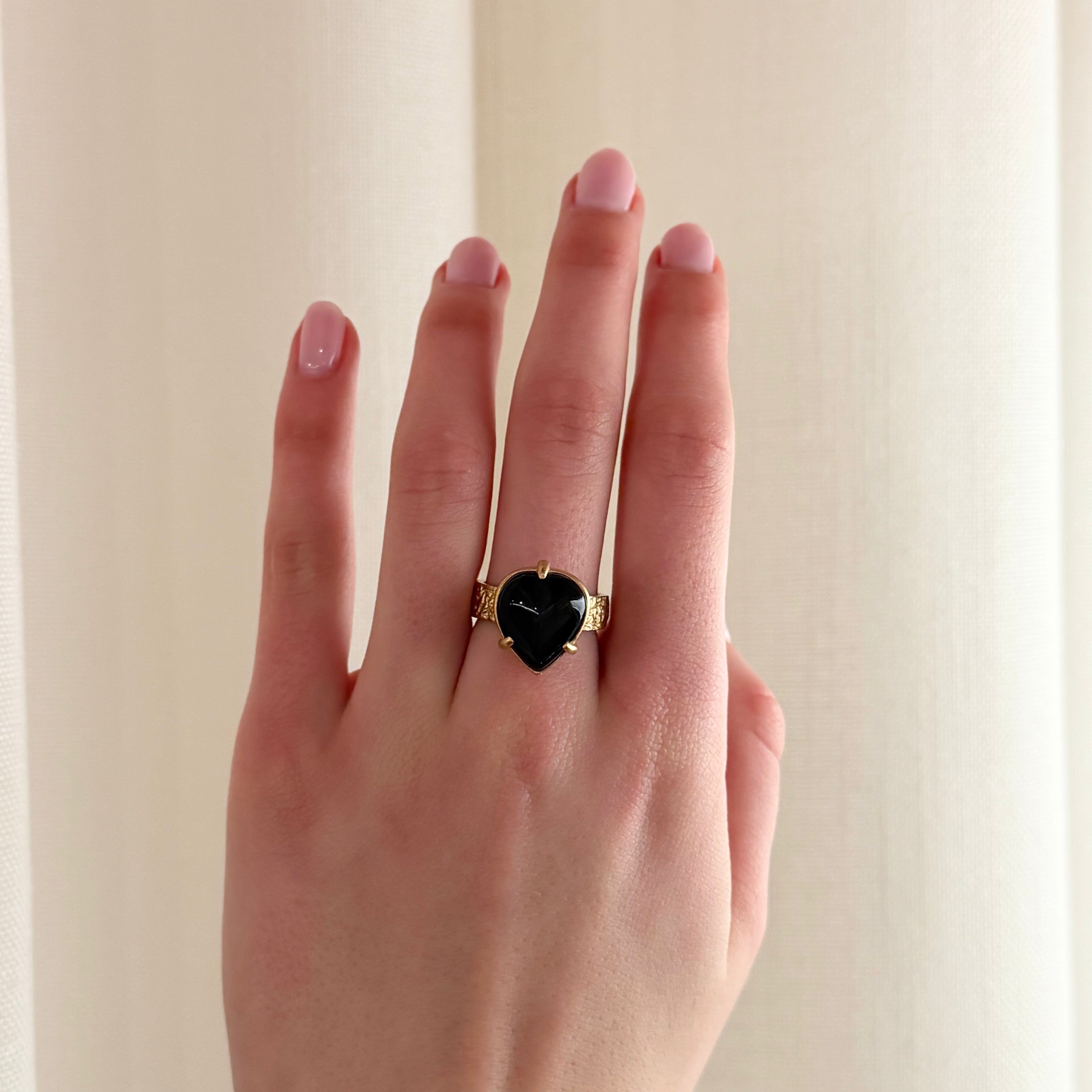 A gold ring with a heart-shaped black gemstone center, worn on a hand with light pink manicured nails, set against a soft beige curtain background.