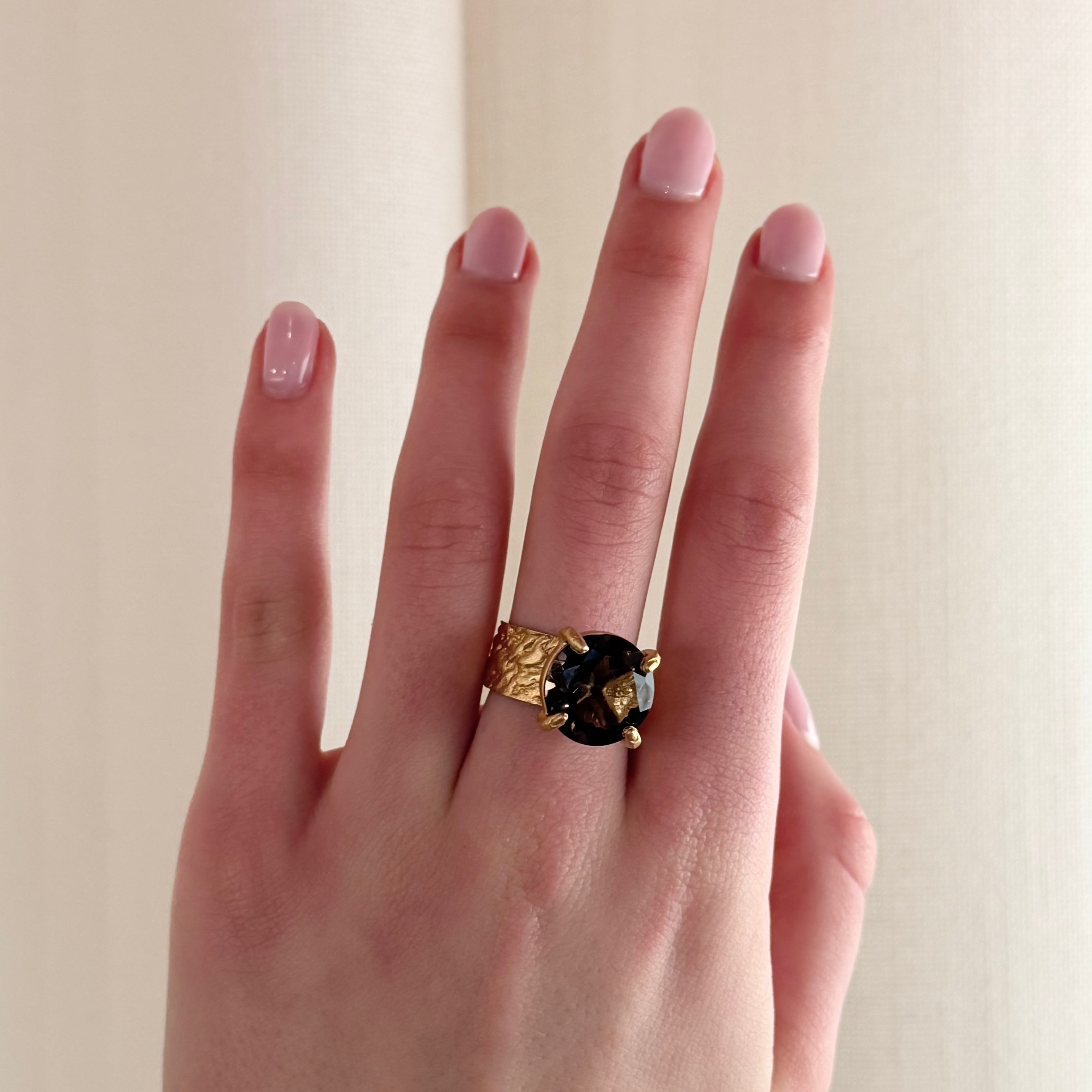 A close-up of a hand wearing a textured gold ring with a wide band, and round brown stone with four claws, set against a soft beige curtain background.