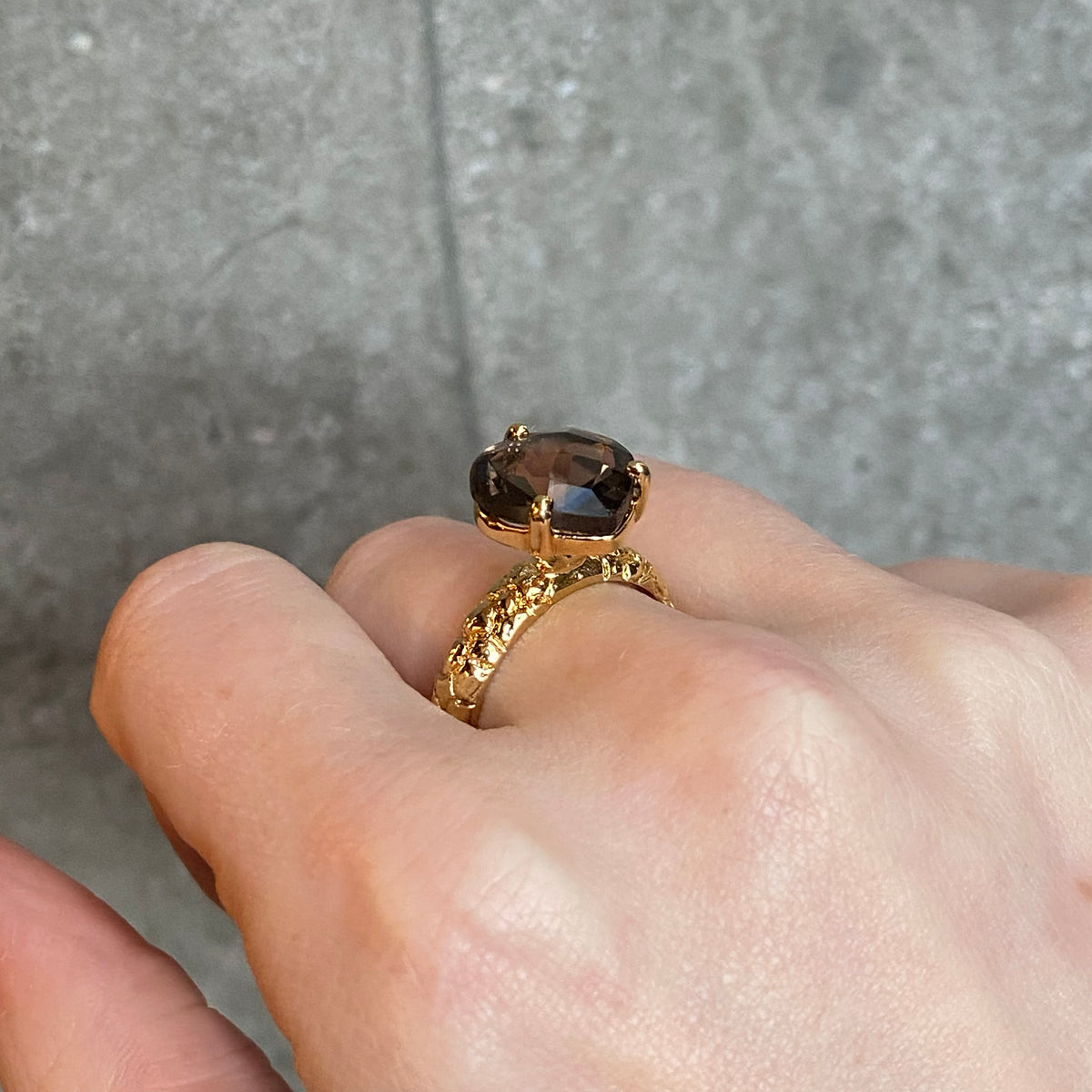 A textured gold ring with a large brown gemstone set within three claws, worn on a person's finger against a grey concrete background.