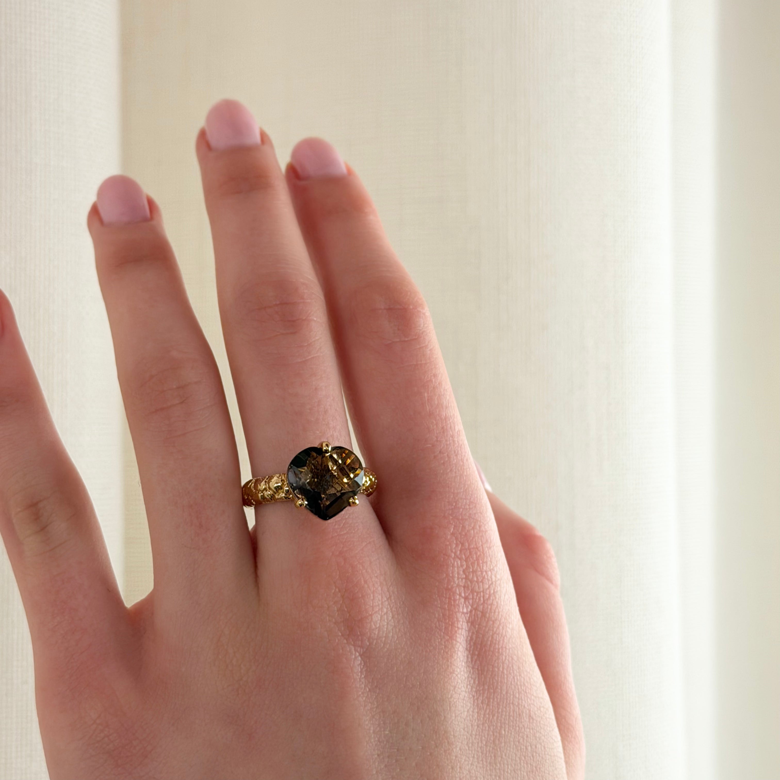 A close-up of a hand wearing a textured gold ring with a dark brown heart-shaped gemstone, set against a soft, off-white curtain background.