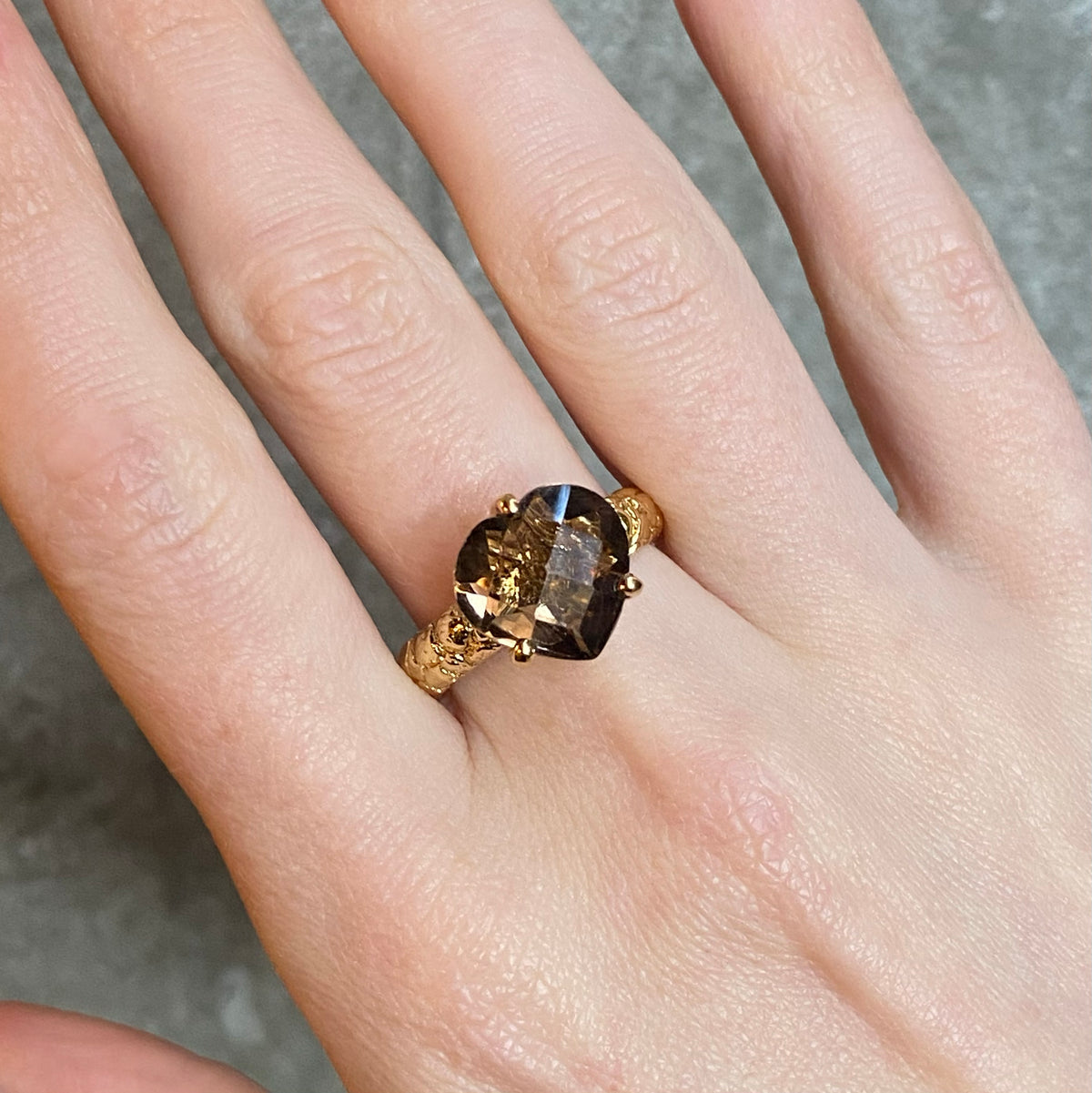A close-up of a hand wearing a textured gold ring with a large heart-shaped brown gemstone held in three claws.