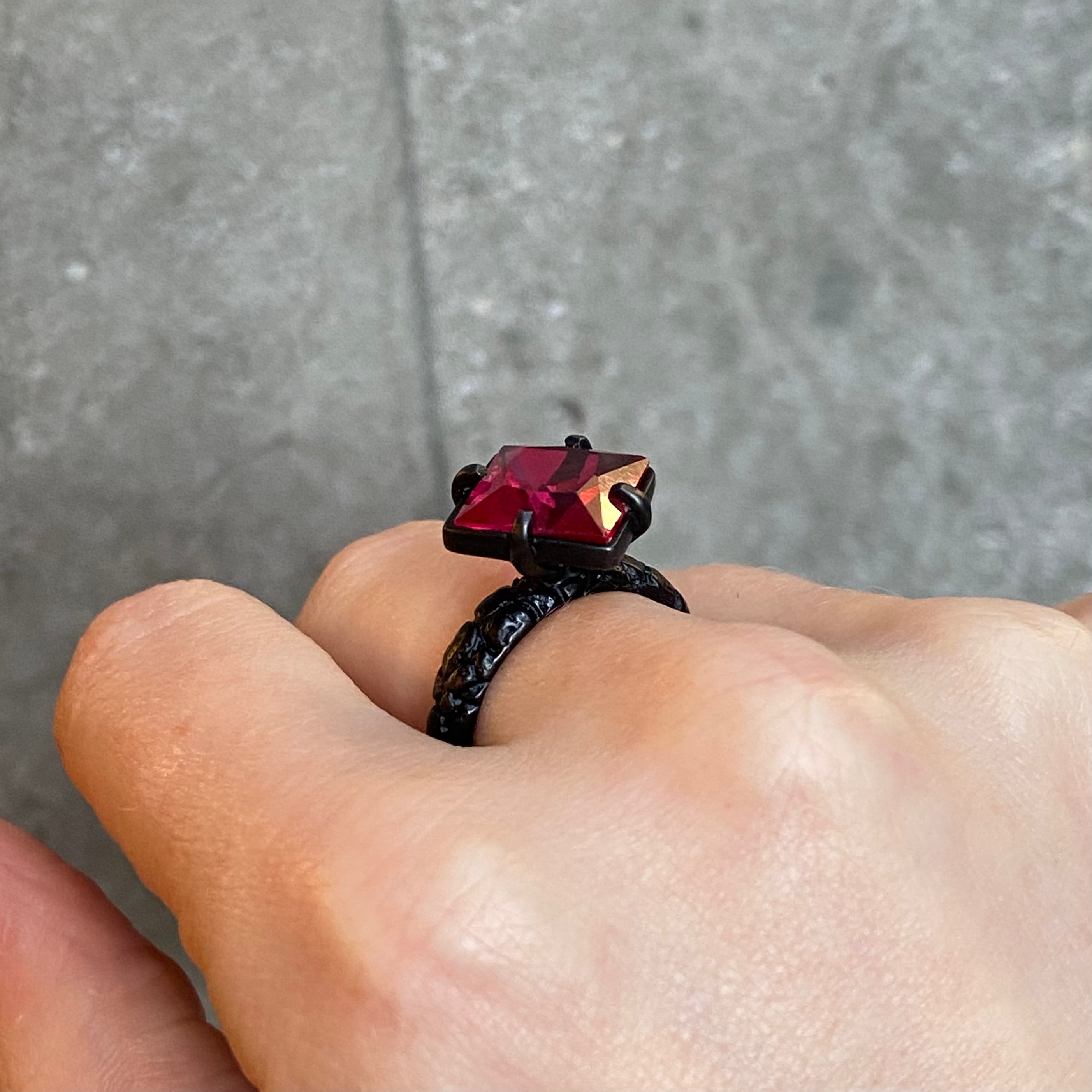 A close-up of a hand wearing a textured black ring with a large, faceted red gemstone set in four claws, against a grey concrete background.