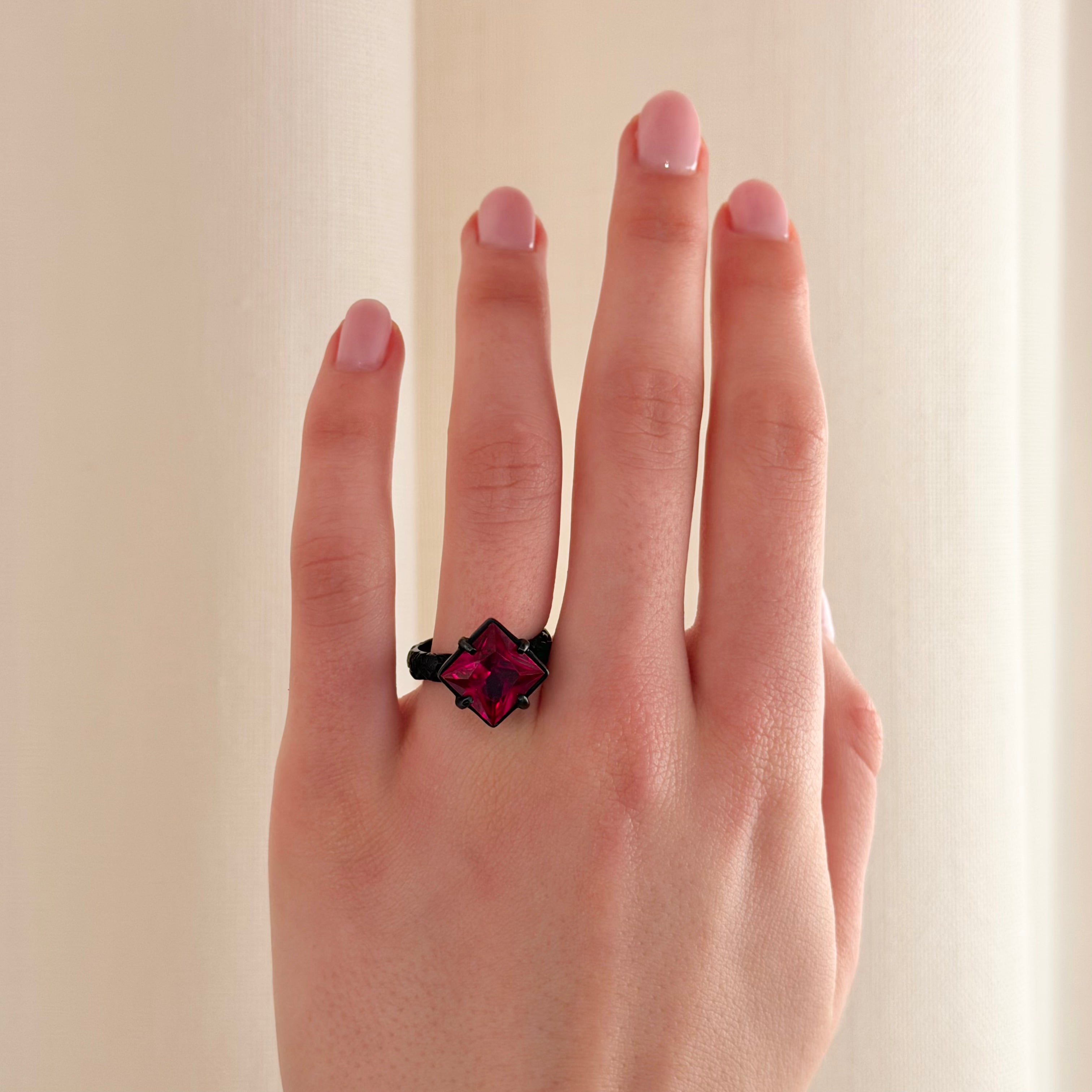 A close-up of a hand wearing a ring with a black band and a prominent deep pink-red faceted gemstone in a claw setting, photographed against a soft cream-coloured curtain background.