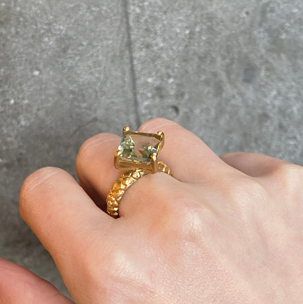 A gold ring with a square-cut pale green gemstone set in four claws. The band is rounded with lots of texture. Worn on a person's finger against a grey concrete background.