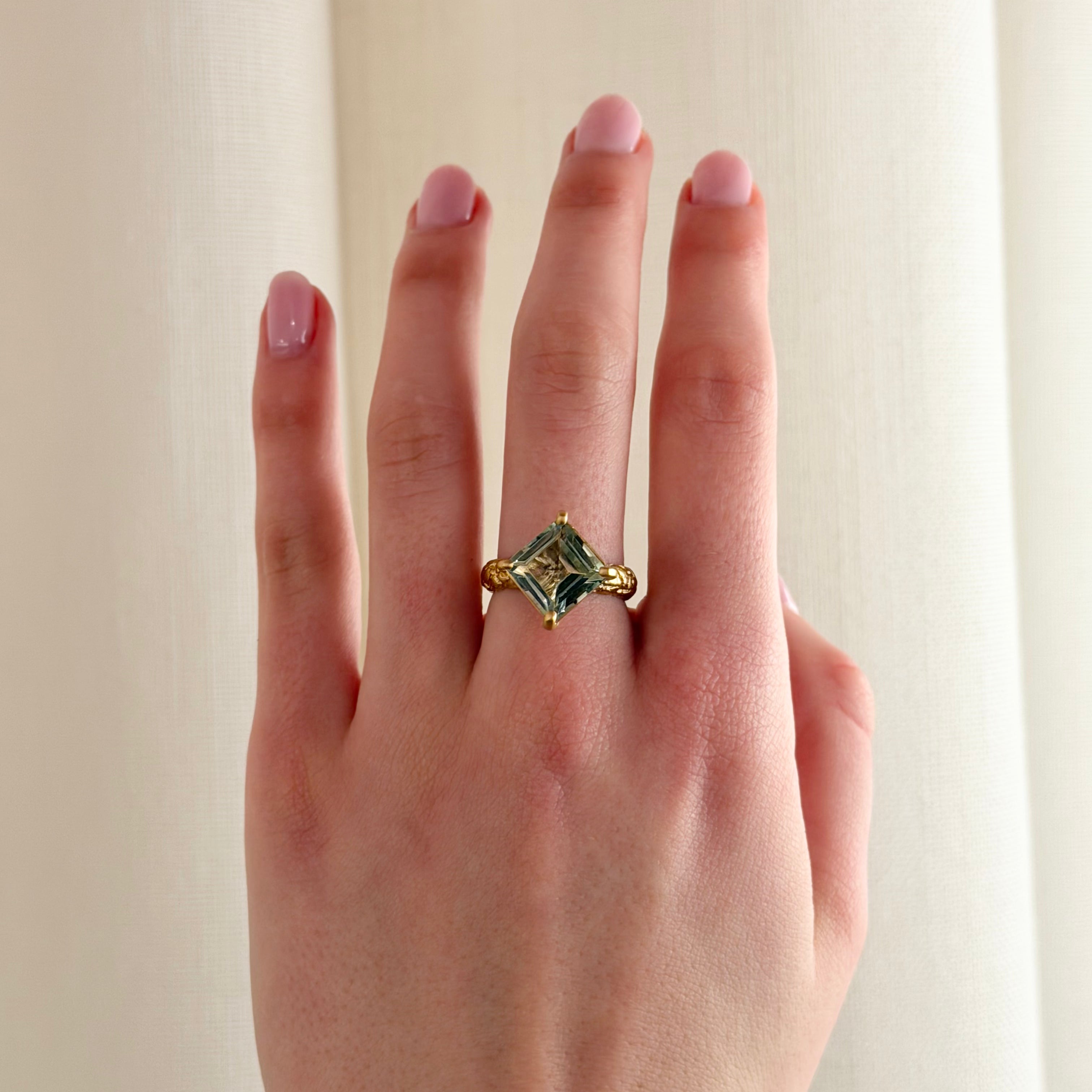 A close-up of a hand wearing a bold gold cocktail ring with a square green gemstone, set against a soft cream-colored curtain background.