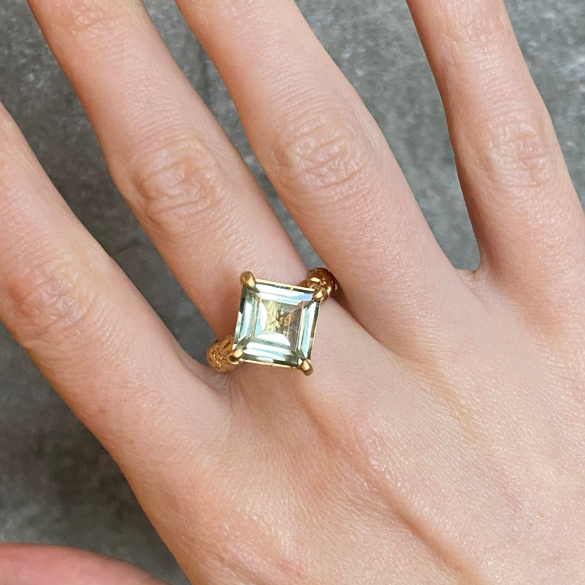 A gold ring with a square-cut pale blue green gemstone worn on a person's finger, shown in a close-up shot against a grey textured background.