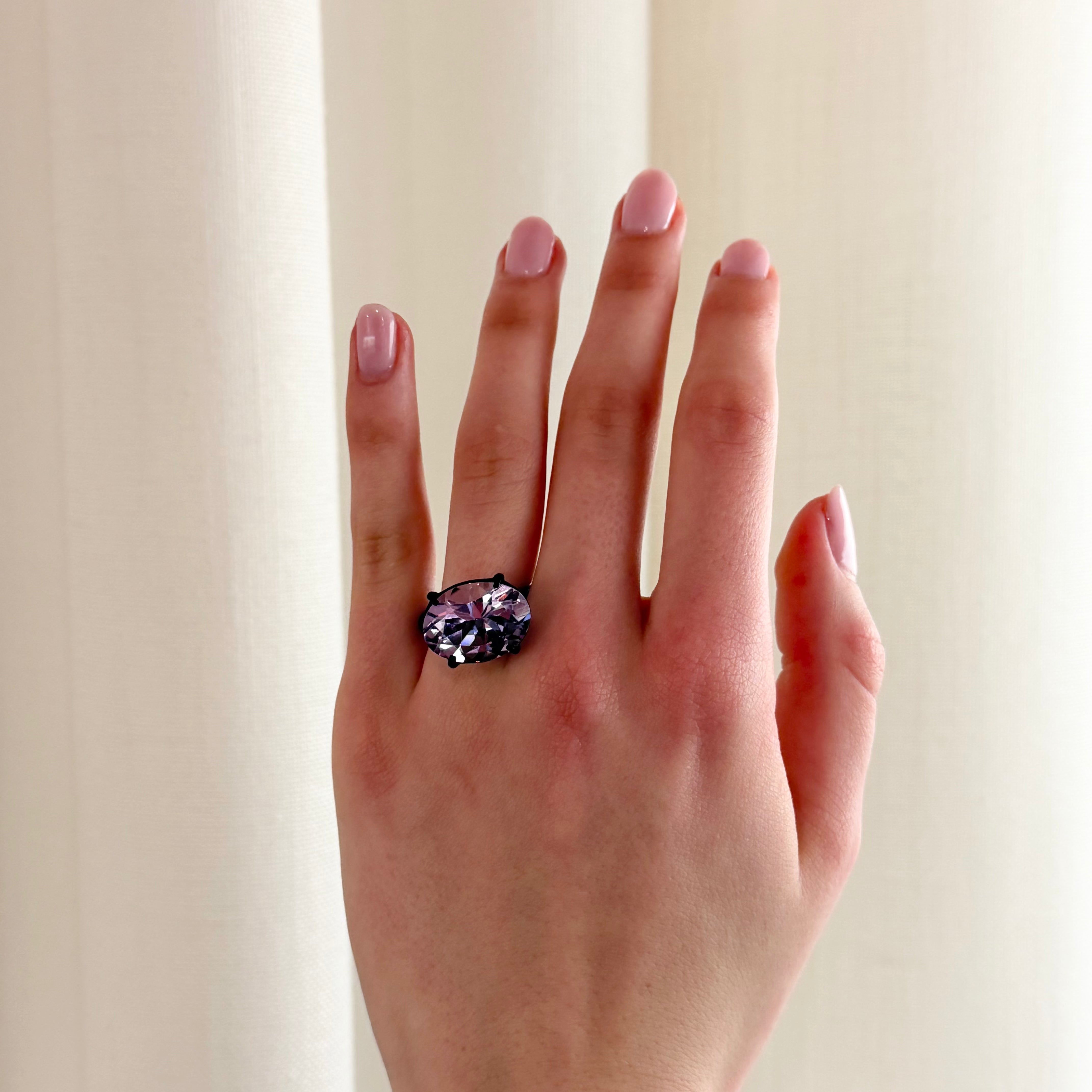 A hand wearing a large cocktail ring with a sparkling oval purple gemstone, set in four dark claws, against a soft cream-coloured curtain background.