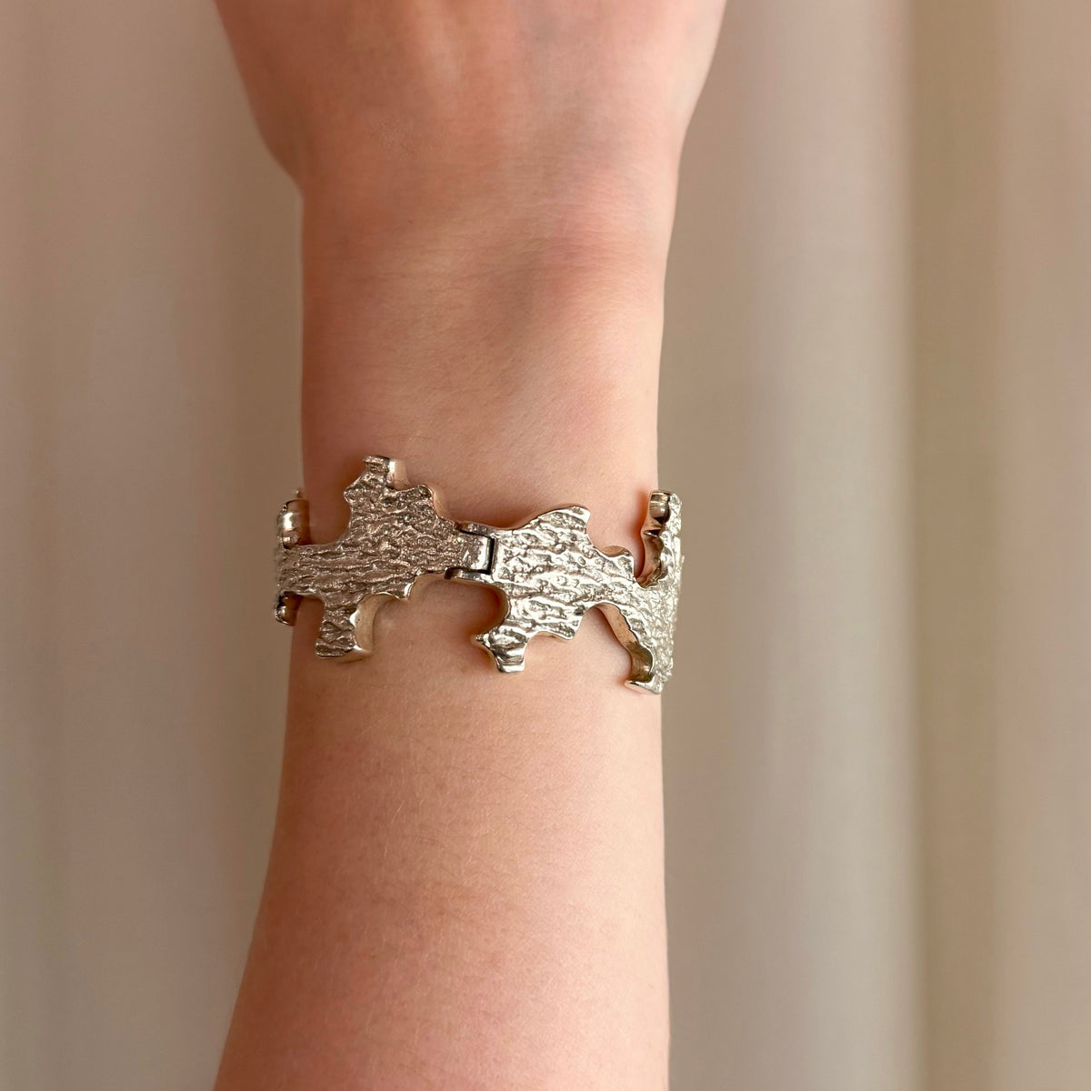 A close-up of a person's wrist wearing an organic textured silver cuff bracelet with irregular cut-out edges, and a hinge mechanism down the middle, set against a soft beige curtain background.