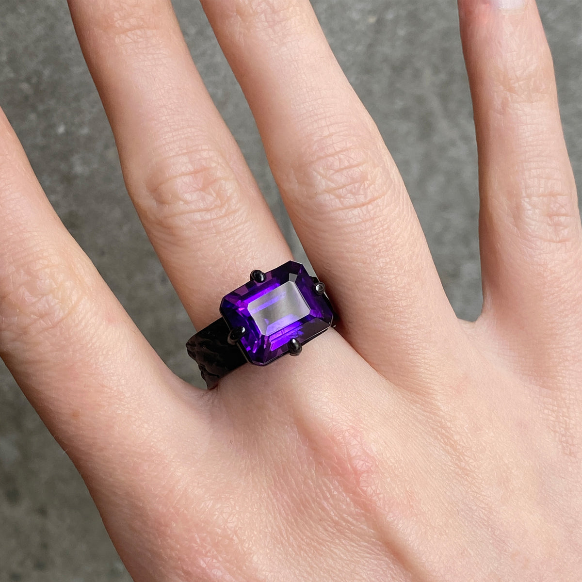 A close-up of a hand wearing a ring with a rectangular purple gemstone in a black band.