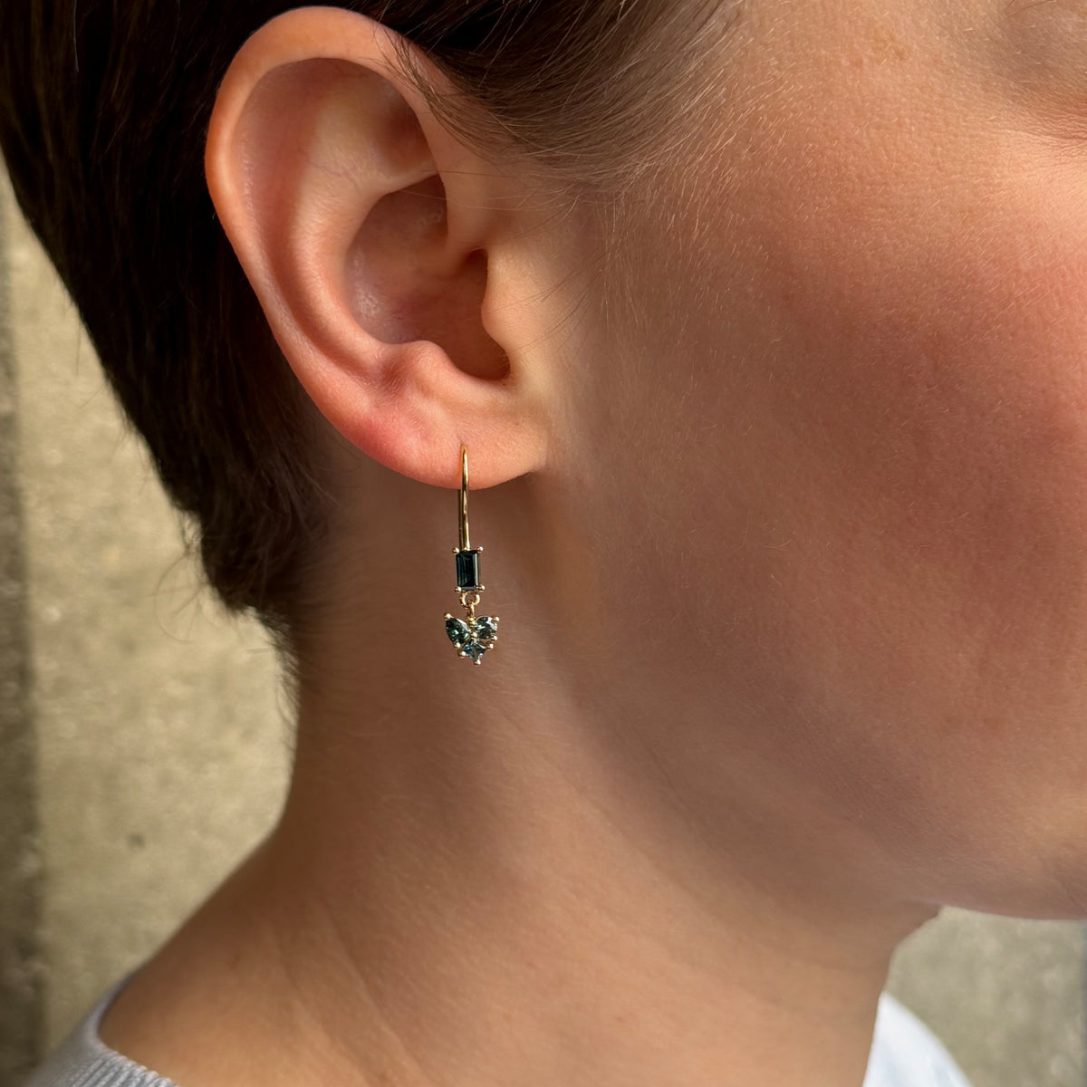 A close up side profile of a person wearing a delicate gold drop earring with a rectangular blue/ green tone and a  cluster of blue-green stones below