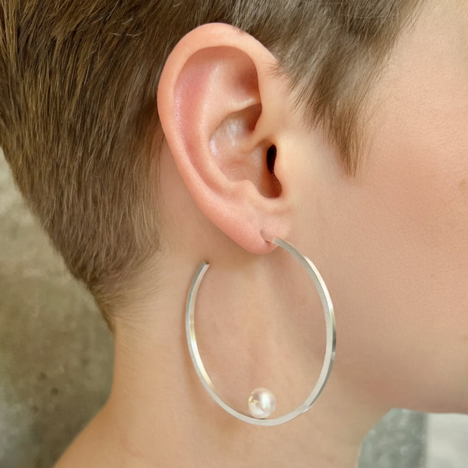 Silver hoop earring with a pearl on an ear against a neutral background