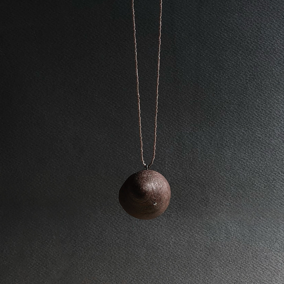 A dark brown textured spherical pendant necklace hanging from a thin beaded cord against a dark grey textured background.