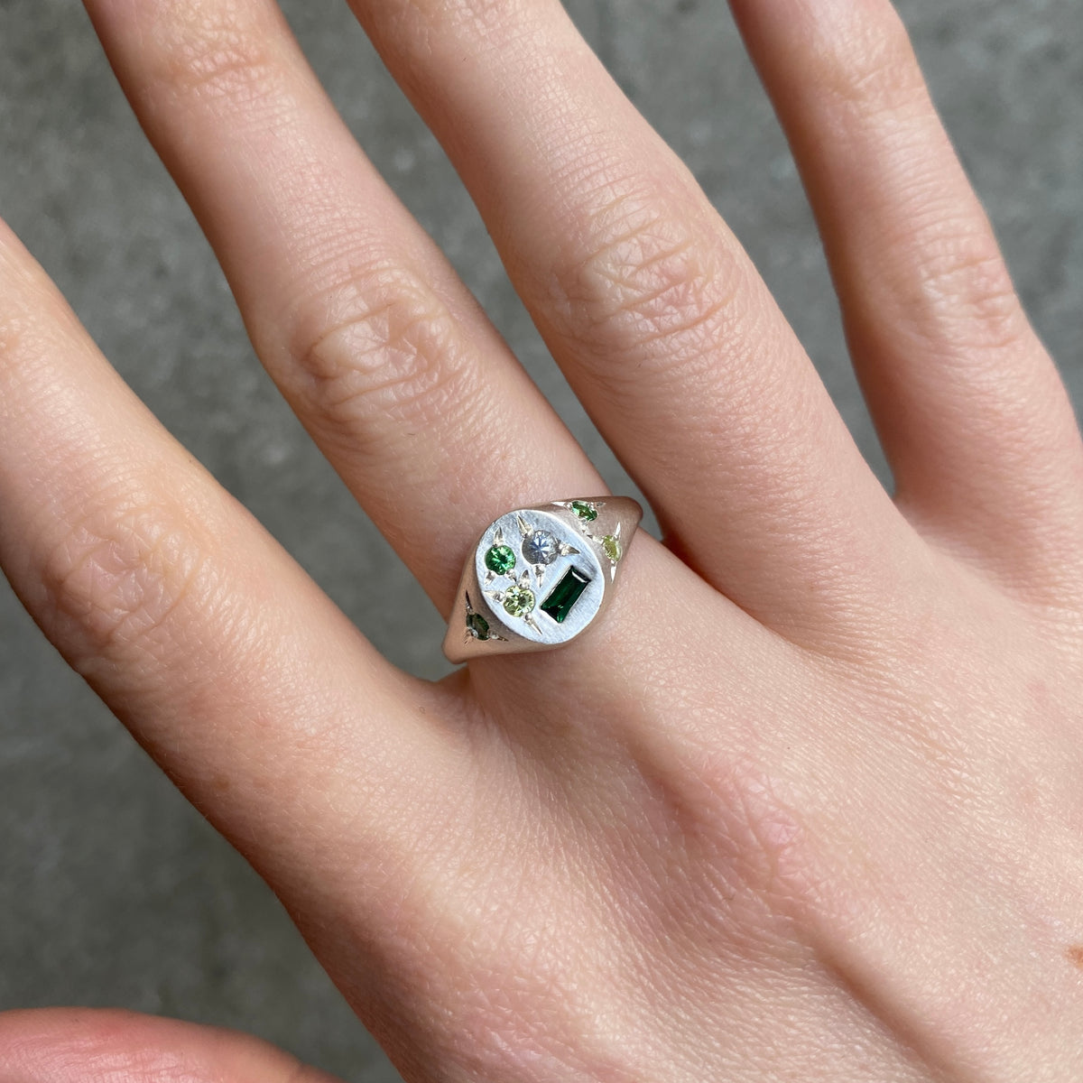 A silver signet ring with a mix of  green and white gemstones, shown worn on a person's ring finger 