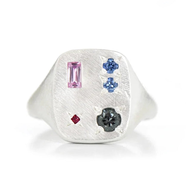 A rectangle silver signet ring featuring a textured flat face with multiple gemstones: a pink baguette, two blue round-cut stones, a small red ruby, and a larger grey round-cut stone.