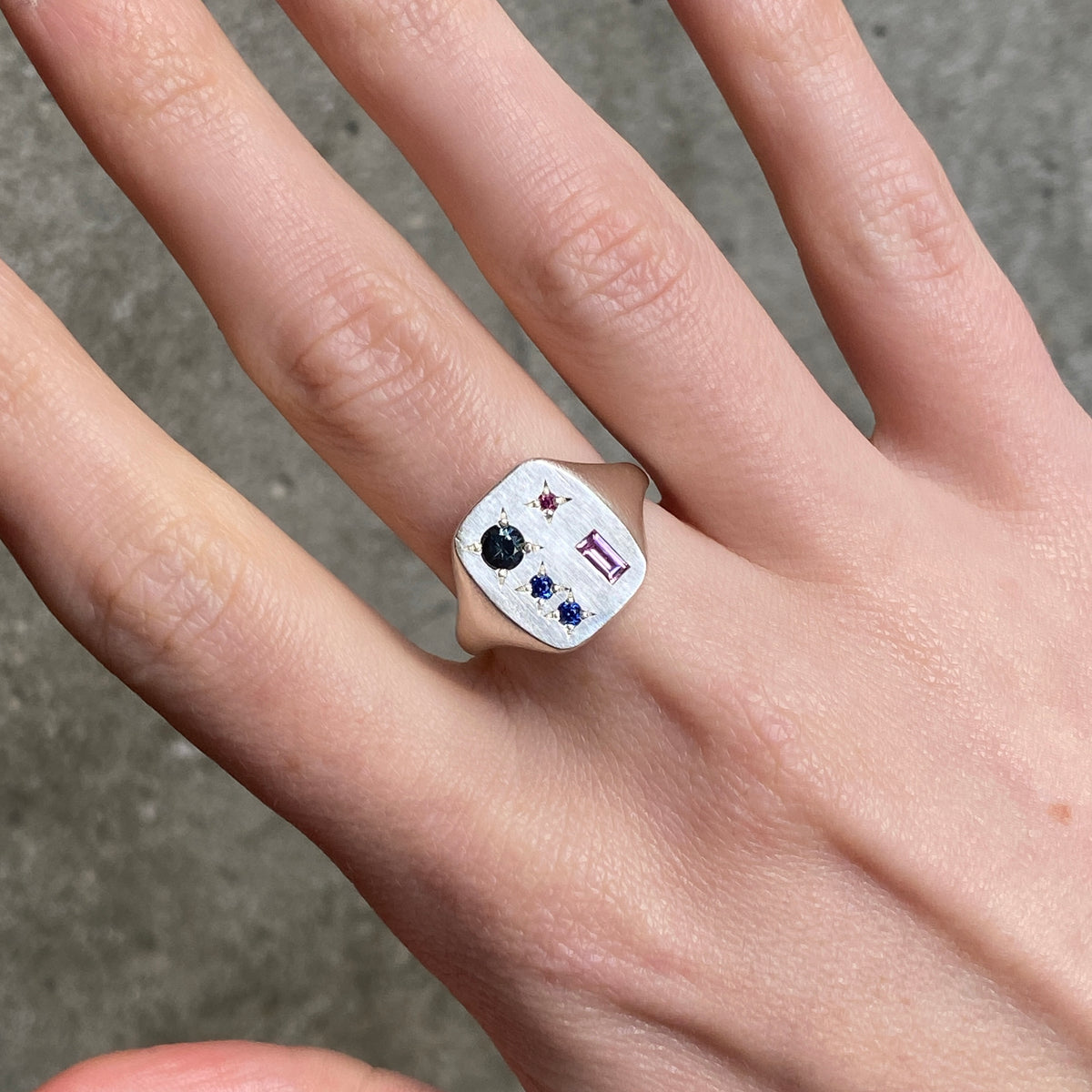 A close up of a hand wearing a square silver ring with grey, blue, red, and pink gemstones