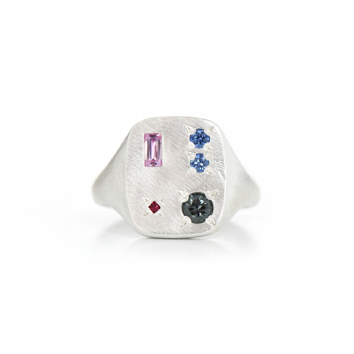 A silver signet ring featuring a rectangular face with multiple coloured gemstones, including a pink baguette, two blue round stones, a small red diamond, and a dark grey stone, set against a plain white background.
