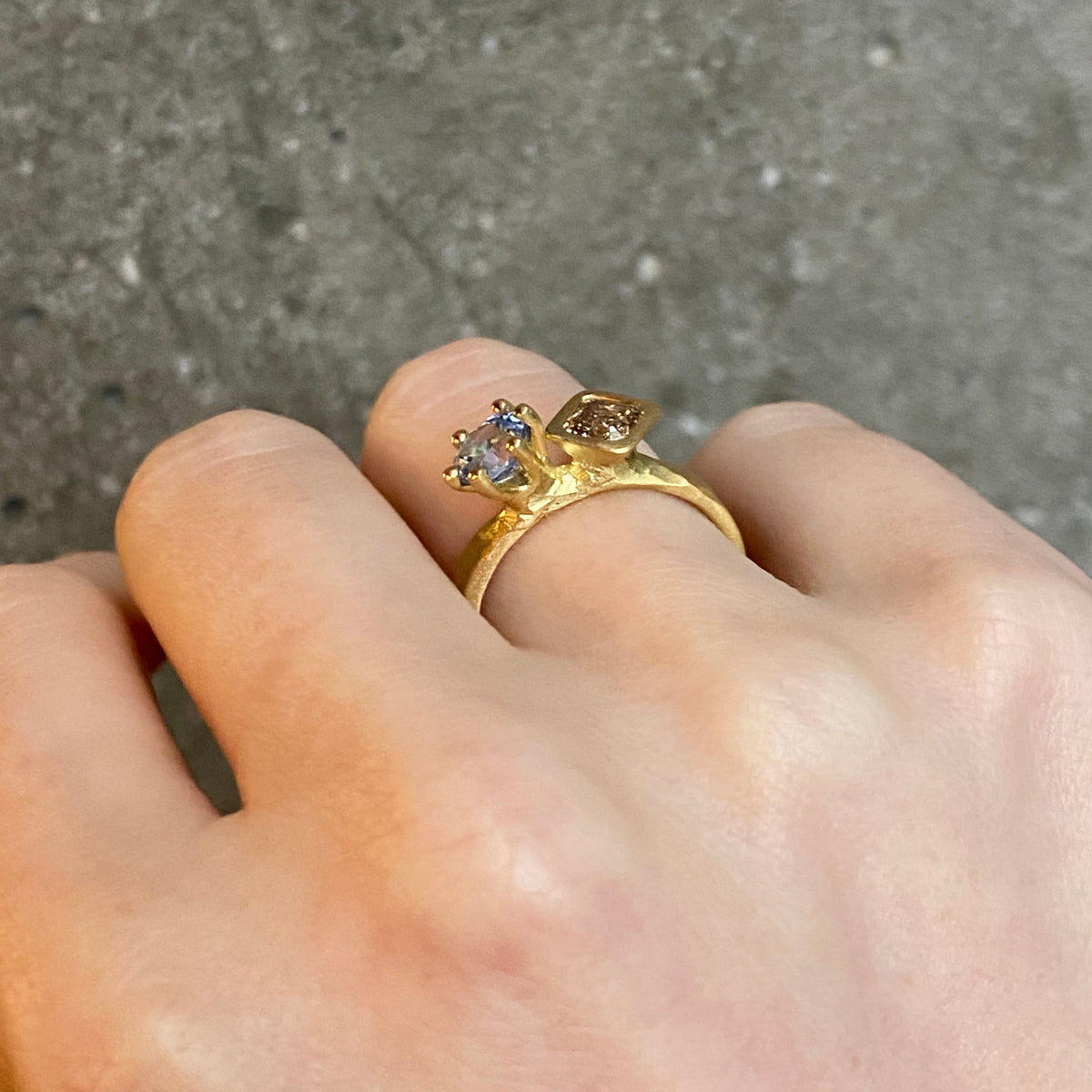 A gold ring with two gemstones of differing shape worn on a person's finger, photographed against a textured gray concrete background.