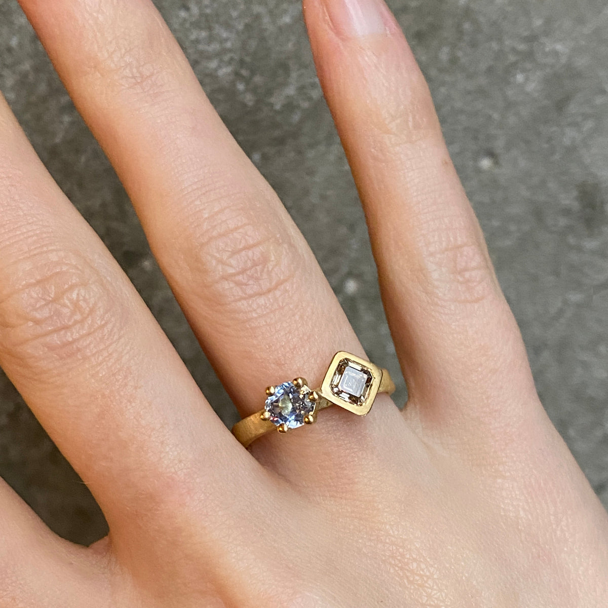 A gold ring with two gemstones of differnt shapes worn on a person's finger, photographed against a textured gray concrete background.