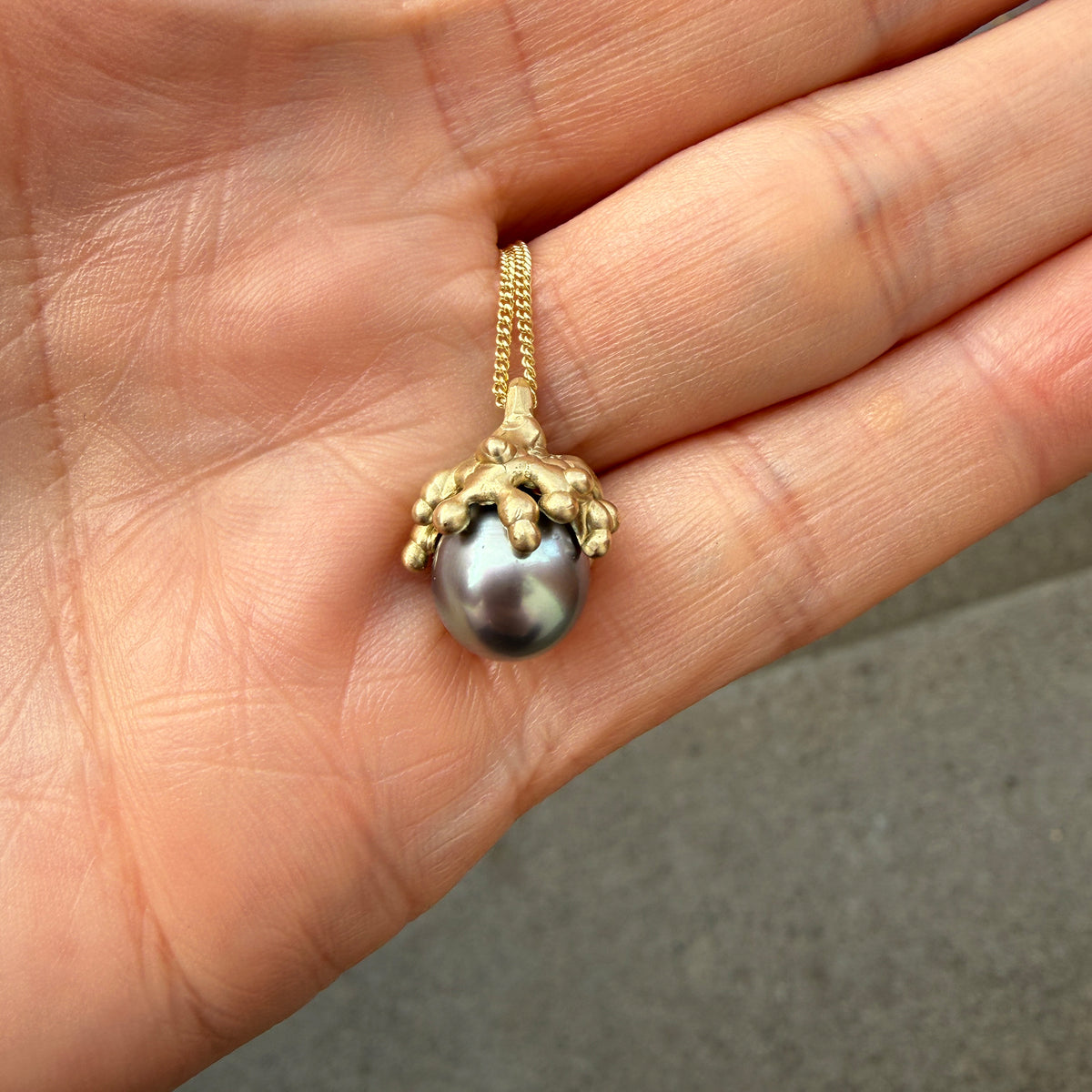 A gold pendant necklace featuring a dark gray pearl set in a textured gold cap, held in a person's hand.
