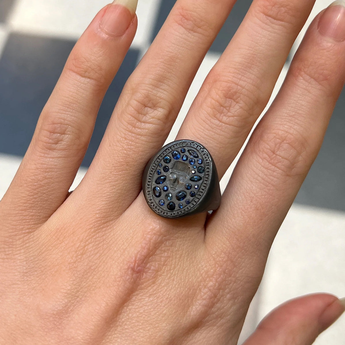 Bold black coloured oval signet ring with an impression of a person's face, surrounded by blue sapphires of mixed cuts and a lightly textured frame. Worn on a hand in front of a chequered white and black background.