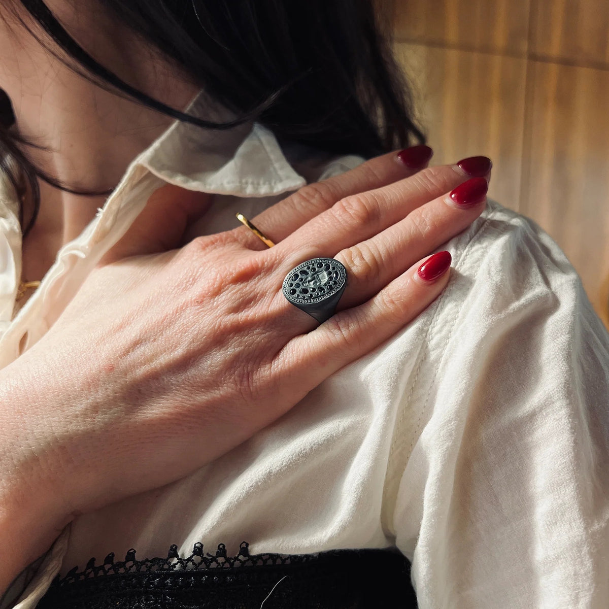 A close-up lifestyle shot of a person wearing a large, round black signet-style ring with an intricate filigree pattern on the top, styled on their middle finger over a white blouse.