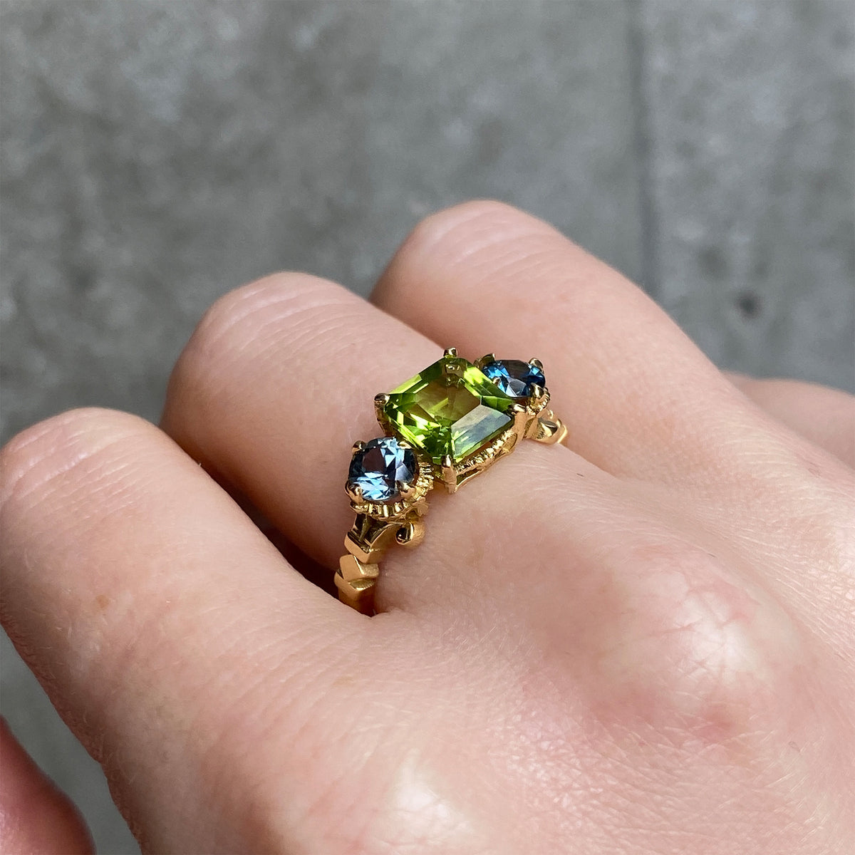 A gold ring with a central peridot and two round blue sapphires on the each side worn on a person's finger.