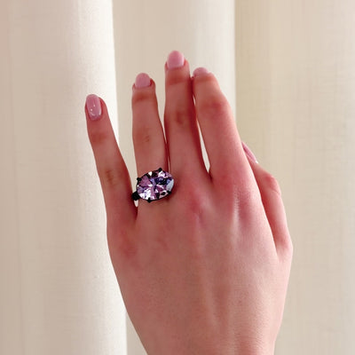 A video of a hand wearing a large cocktail ring with a sparkling oval purple gemstone, set in four dark claws, against a soft cream-coloured curtain background.