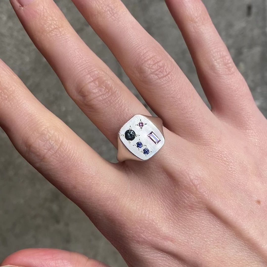 A close up of a hand wearing a square silver ring with grey, blue, red, and pink gemstones