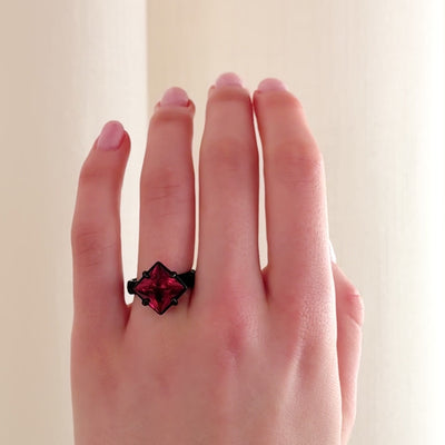 A close-up video of a hand wearing a ring with a black band and a prominent deep pink-red faceted gemstone in a four claw setting, photographed against a soft cream-coloured curtain background.