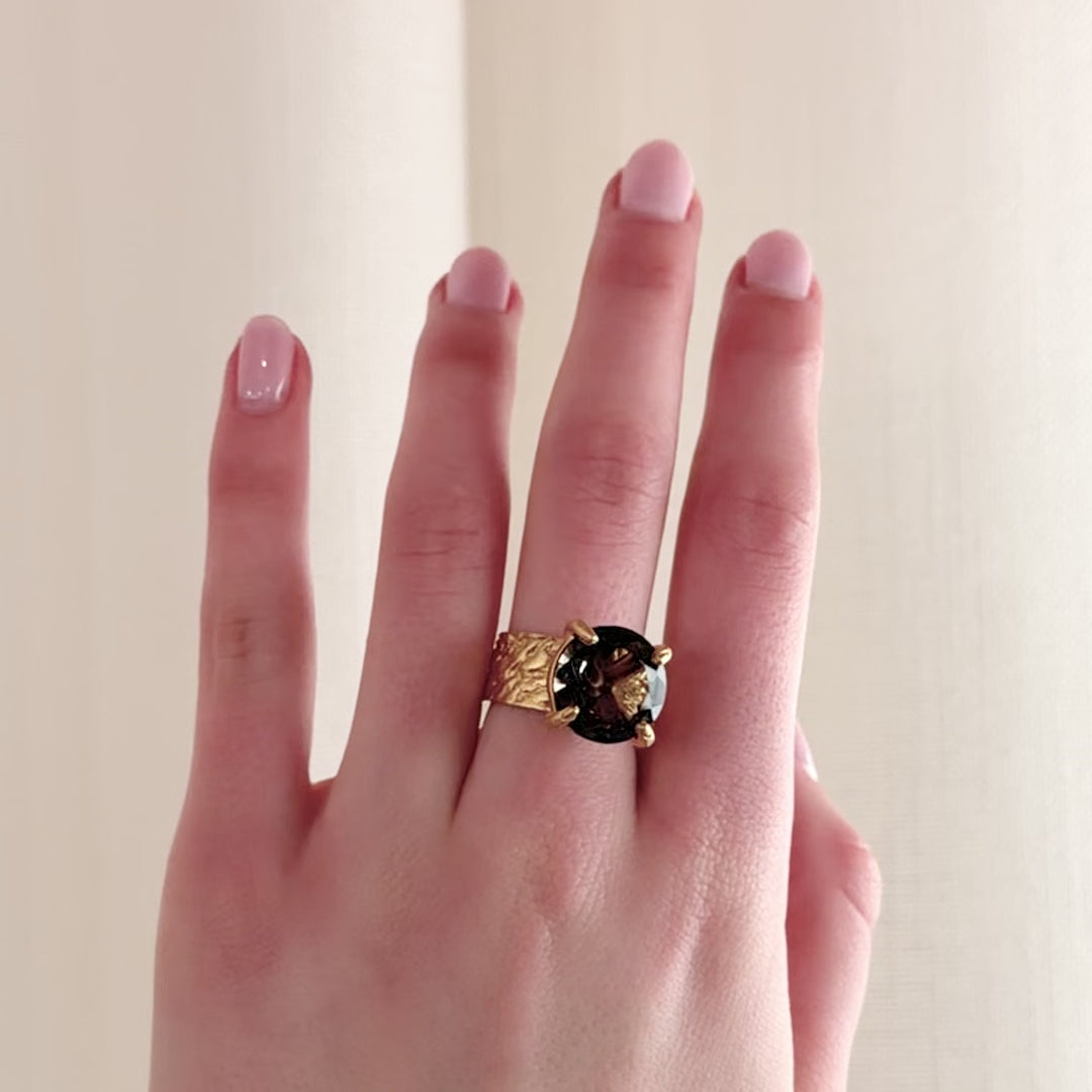 A close-up video of a hand wearing a textured gold ring with a wide band, and round brown stone with four claws, set against a soft beige curtain background.