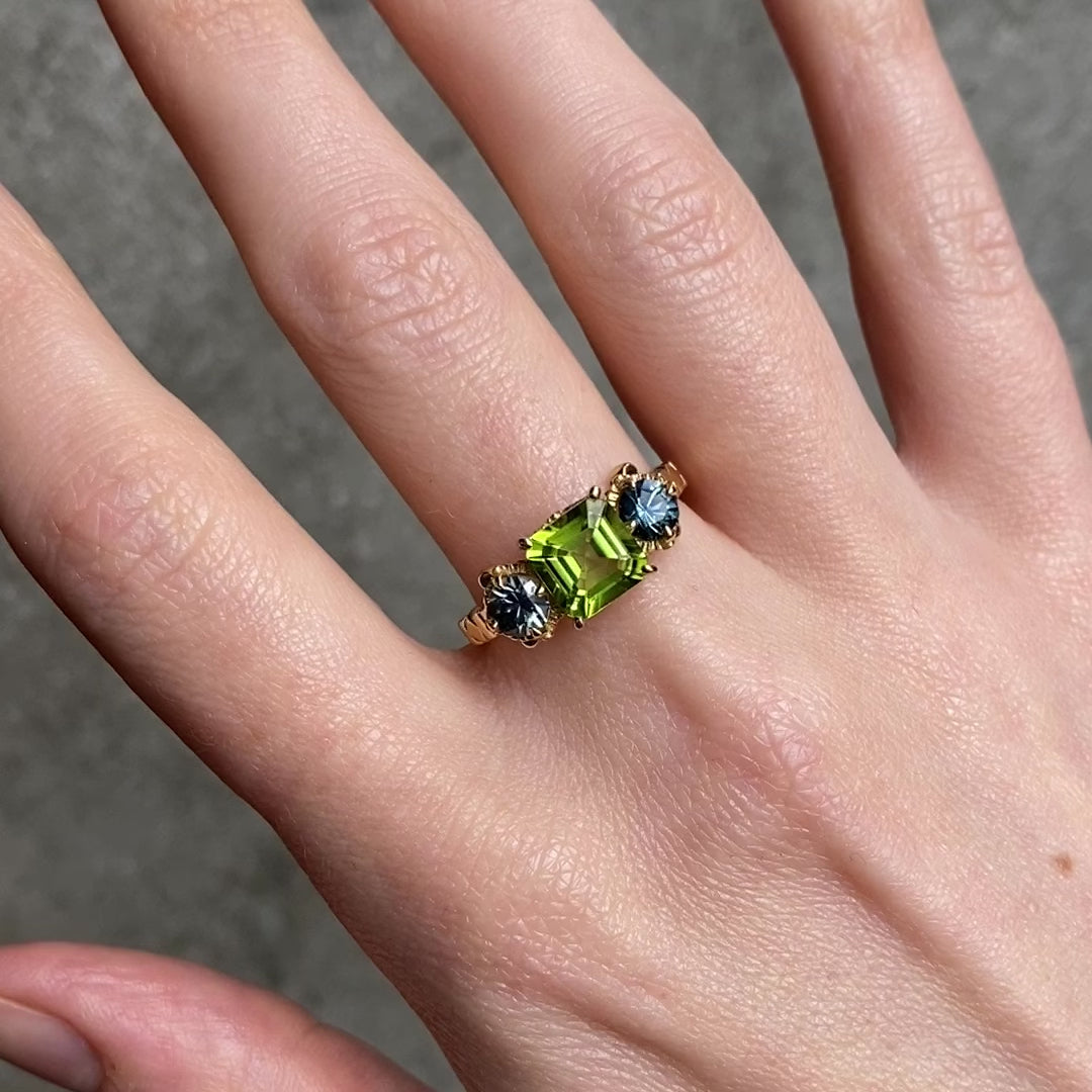 A gold ring with a large green peridot and two blue sapphires, shown worn on a person's finger against a gray concrete background.