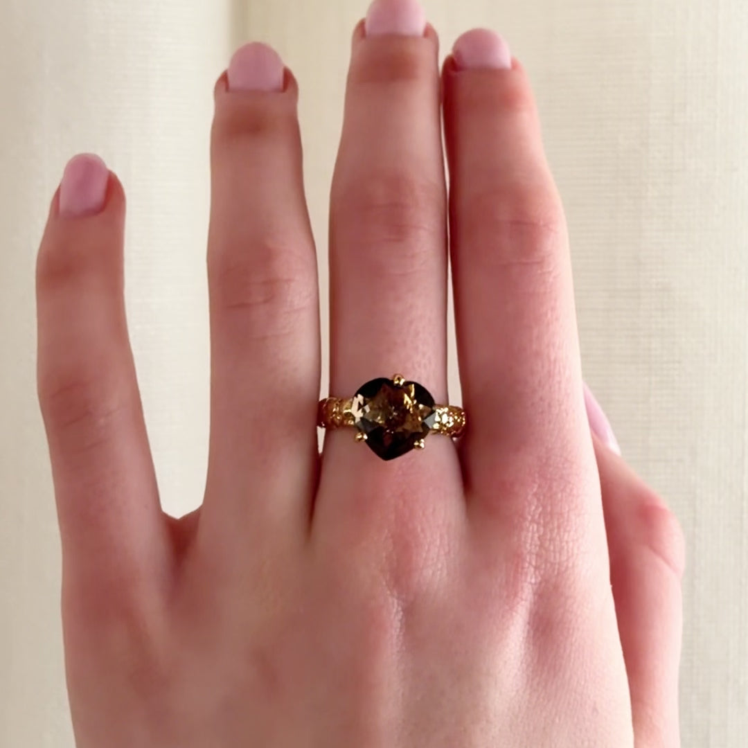 A close-up video of a hand wearing a textured gold ring with a dark brown heart-shaped gemstone, set against a soft, off-white curtain background.