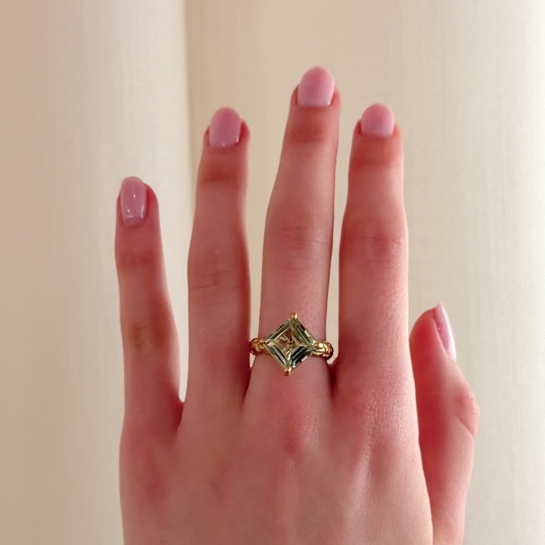 A close-up video of a hand wearing a bold gold cocktail ring with a square pale green gemstone, set against a soft cream-colored curtain background.
